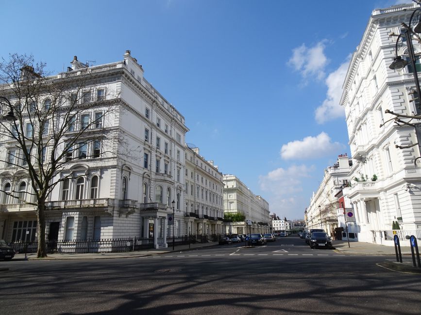 A wide view of a street in an affluent neighbourhood with white, ornate terraced townhouses featuring large windows, decorative cornices, and small balconies. The street has parked cars along both sides and a footpath with a black wrought iron fence. The sky is clear with some clouds, and a leafless tree is visible in the foreground. The image captures the exterior environment of a house removal process, suggesting the location is near Old Brompton Road in London, and is relevant for furniture transport and home relocation services provided by Brompton Removals, with the focus on the urban setting and building facades in the context of house moving logistics.