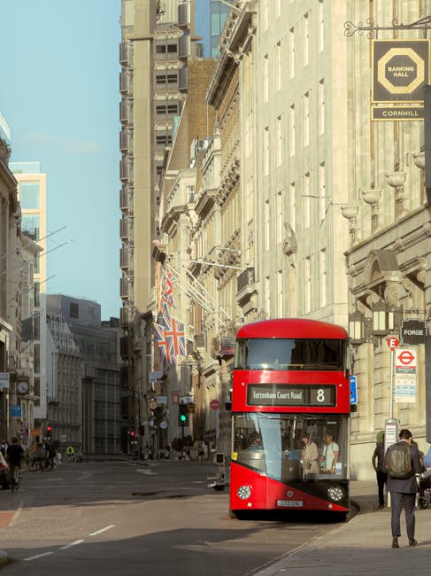 A red double-decker bus numbered 8 is approaching an intersection on a busy street in central London, with tall historic and modern buildings lining the roadway. The buildings feature ornate stone facades, large windows, and decorative architectural details, with some displaying Union Jack flags and street signage. Pedestrians are walking along the pavement, some carrying bags or backpacks, while others wait near the crosswalk. Traffic lights are visible, and a black and yellow banking hall sign is mounted on one building. The street appears to be part of a central commercial or financial district, with a mixture of foot and vehicle traffic. Brompton Removals' vehicle and moving equipment are not present in this street scene, as the image captures the urban environment rather than the loading process or home relocation activities. The setting is well-lit with clear daytime weather, emphasizing the city's characteristic architecture and lively atmosphere.