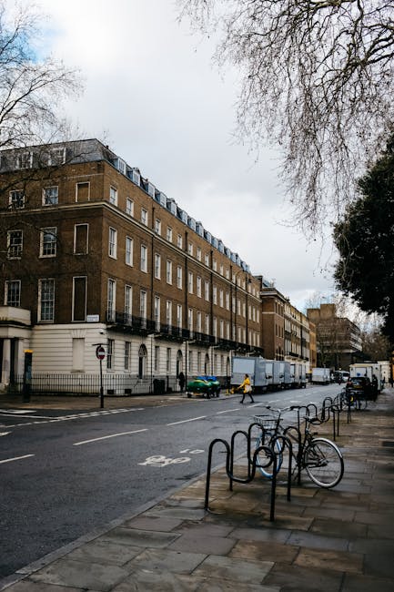 A wide view of a street in an affluent neighbourhood with white, ornate terraced townhouses featuring large windows, decorative cornices, and small balconies. The street has parked cars along both sides and a footpath with a black wrought iron fence. The sky is clear with some clouds, and a leafless tree is visible in the foreground. The image captures the exterior environment of a house removal process, suggesting the location is near Old Brompton Road in London, and is relevant for furniture transport and home relocation services provided by Brompton Removals, with the focus on the urban setting and building facades in the context of house moving logistics.